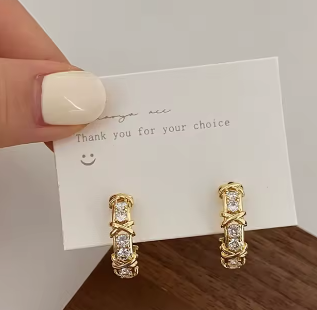 Aros huggies dorados con zirconia AAA diseño cruzado X-Twirl
Detalle de engaste cruzado en aros dorados con brillo
Aretes huggie chapados en oro 18K sobre superficie de madera
Modelo usando aros huggies dorados tamaño 20 mm