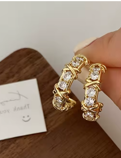 Aros huggies dorados con zirconia AAA diseño cruzado X-Twirl

Detalle de engaste cruzado en aros dorados con brillo

Aretes huggie chapados en oro 18K sobre superficie de madera

Modelo usando aros huggies dorados tamaño 20 mm